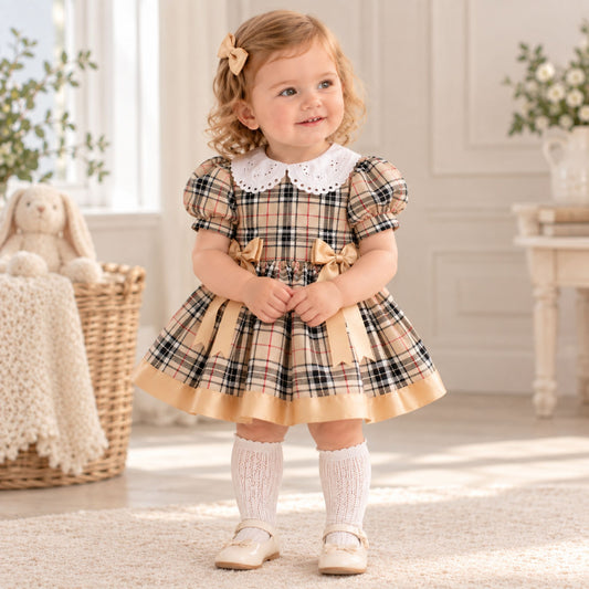 A toddler girl wearing a short-sleeve plaid dress with a white lace Peter Pan collar, cream ruffled hem, and tan bows, standing in a bright, neutral-toned room.