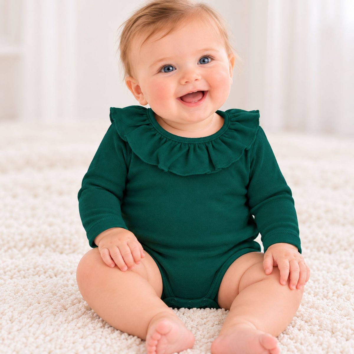 A smiling baby sitting on a white textured rug wearing a dark green long-sleeve romper with a ruffled collar.