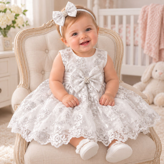 A baby girl wearing a sleeveless white lace dress with a large bow at the chest and a matching bow headband, sitting on a cream upholstered armchair in a softly lit nursery.