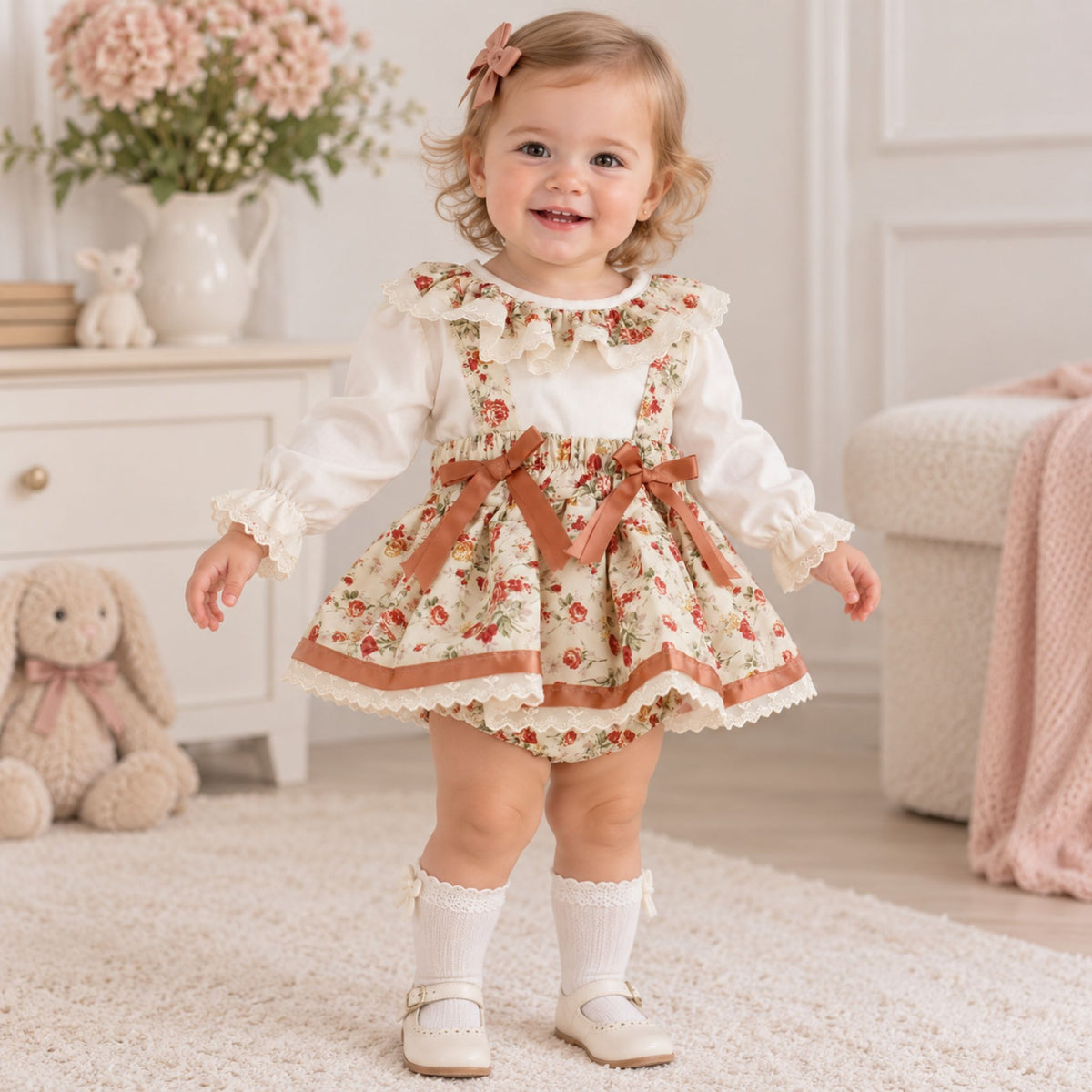 A toddler girl stands on a cream rug in a bright, soft room, wearing a cream long-sleeve blouse under a floral bib-style dress with brown ribbon trim and bows. She has white socks and cream shoes, with a pink bow in her hair.