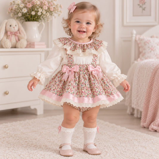 A toddler girl stands in a softly lit nursery wearing a floral baby dress with a white long-sleeve blouse underneath, pink bows, and lace trim. The room includes a white dresser, bunny plush, flowers, and a pink bed.