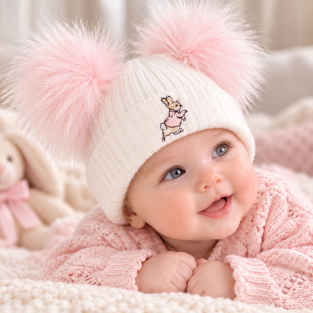 A baby wearing a white ribbed knit beanie hat with two fluffy pink pompoms and a small bunny embroidery, lying on a pink blanket next to a plush toy.