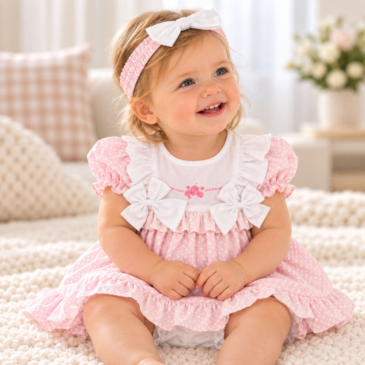 A baby girl wearing a pink and white polka dot dress with white ruffle bows and a matching pink and white polka dot headband, sitting on a soft cream knit blanket in a cozy room.
