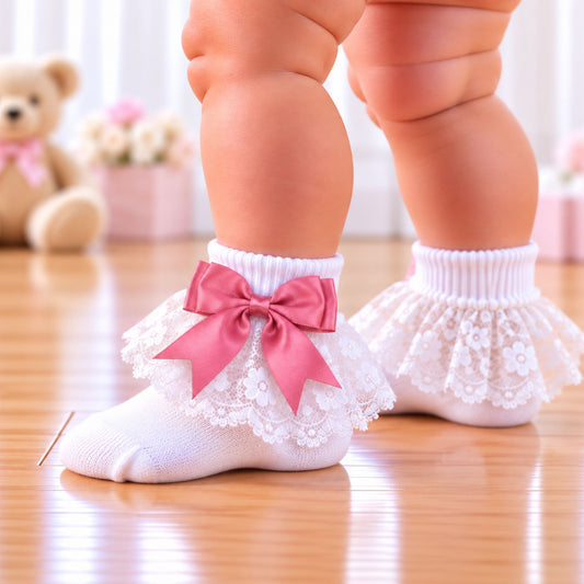 White baby socks with lace ruffles and pink bows, worn on a baby's feet on a wooden floor.