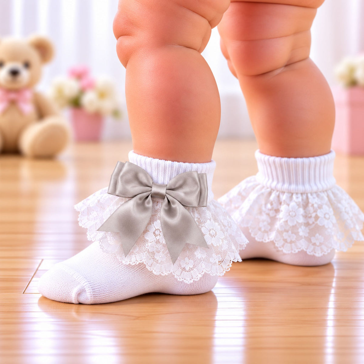White lace baby socks with ruffled trim and a satin bow, worn on a baby's feet on a wooden floor with a blurred teddy bear in the background.