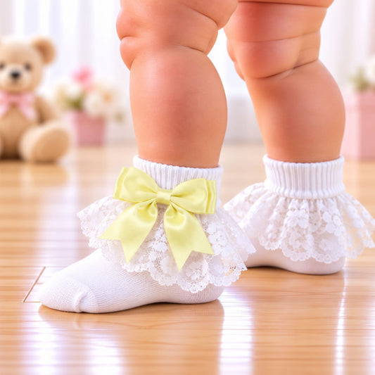 White lace baby socks with ruffled trim and a pale yellow bow, worn on a baby's feet on a wooden floor with a blurred teddy bear in the background.