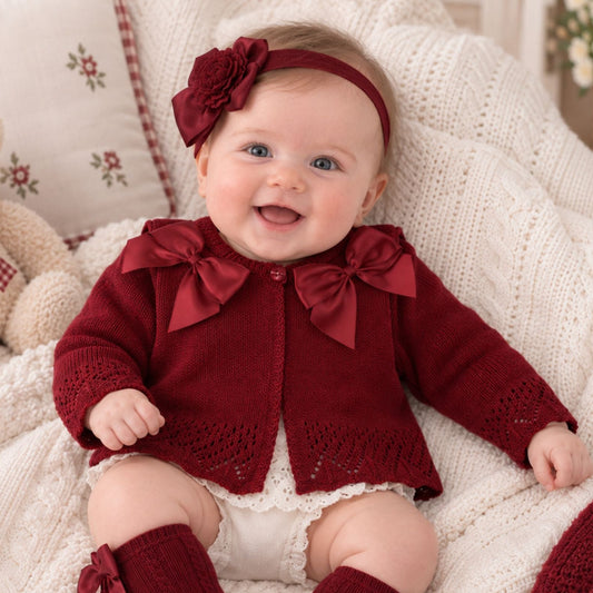 A baby wearing a burgundy knit cardigan with large bows, matching knee-high socks, and a headband, sitting on a white textured blanket next to a teddy bear.