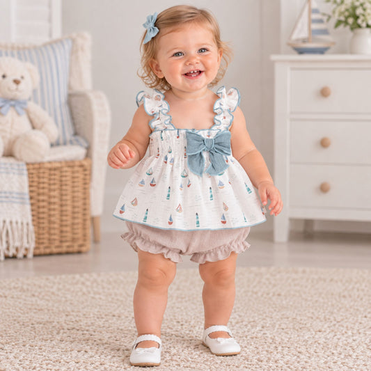 A toddler girl wearing a white sleeveless romper dress with a blue bow and sailboat print, paired with light beige ruffled bloomers and white Mary Jane shoes, standing in a bright nursery setting.