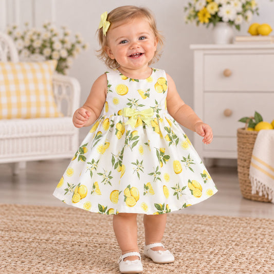A toddler girl wearing a sleeveless white dress with a yellow lemon and green leaf print, standing in a bright, decorated room. She has a yellow bow in her hair and is wearing white Mary Jane shoes.