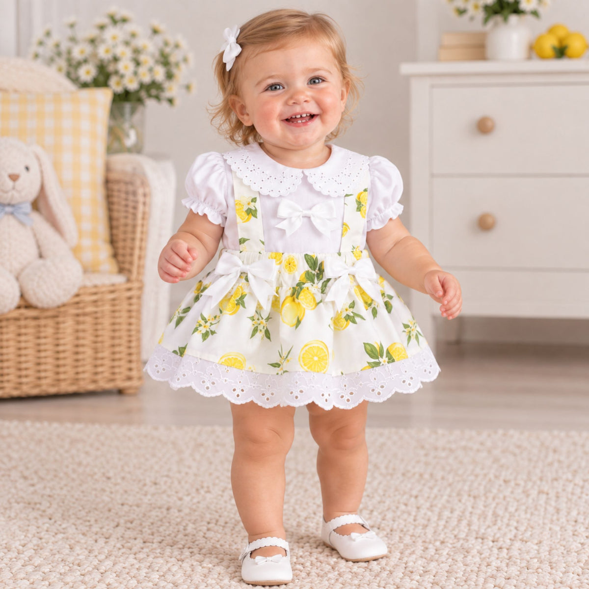 A toddler girl wearing a white short-sleeve dress with a yellow lemon print and white lace trim, standing on a beige rug in a bright nursery setting.