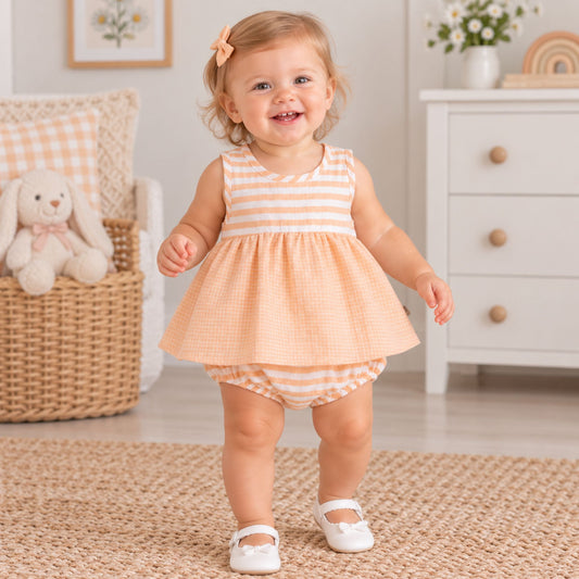 A smiling toddler girl wearing a peach and white gingham sleeveless dress with matching striped bloomers and white Mary Jane shoes, standing on a woven rug in a bright nursery setting.