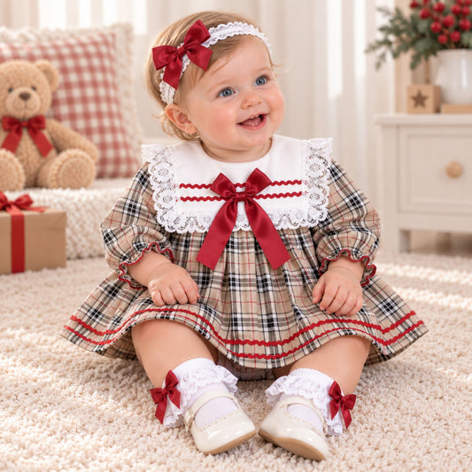A baby girl wearing a Christmas-themed plaid dress with a white lace collar and red ribbon bow, sitting on a beige carpet in a cozy room.
