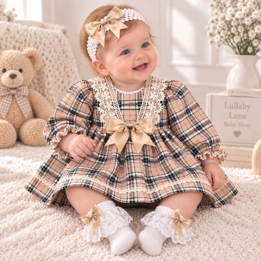 A baby girl sitting on a cream rug wearing a beige plaid dress with white lace trim, a large beige bow at the chest, and matching lace booties with bows. She is wearing a white headband with a beige bow.
