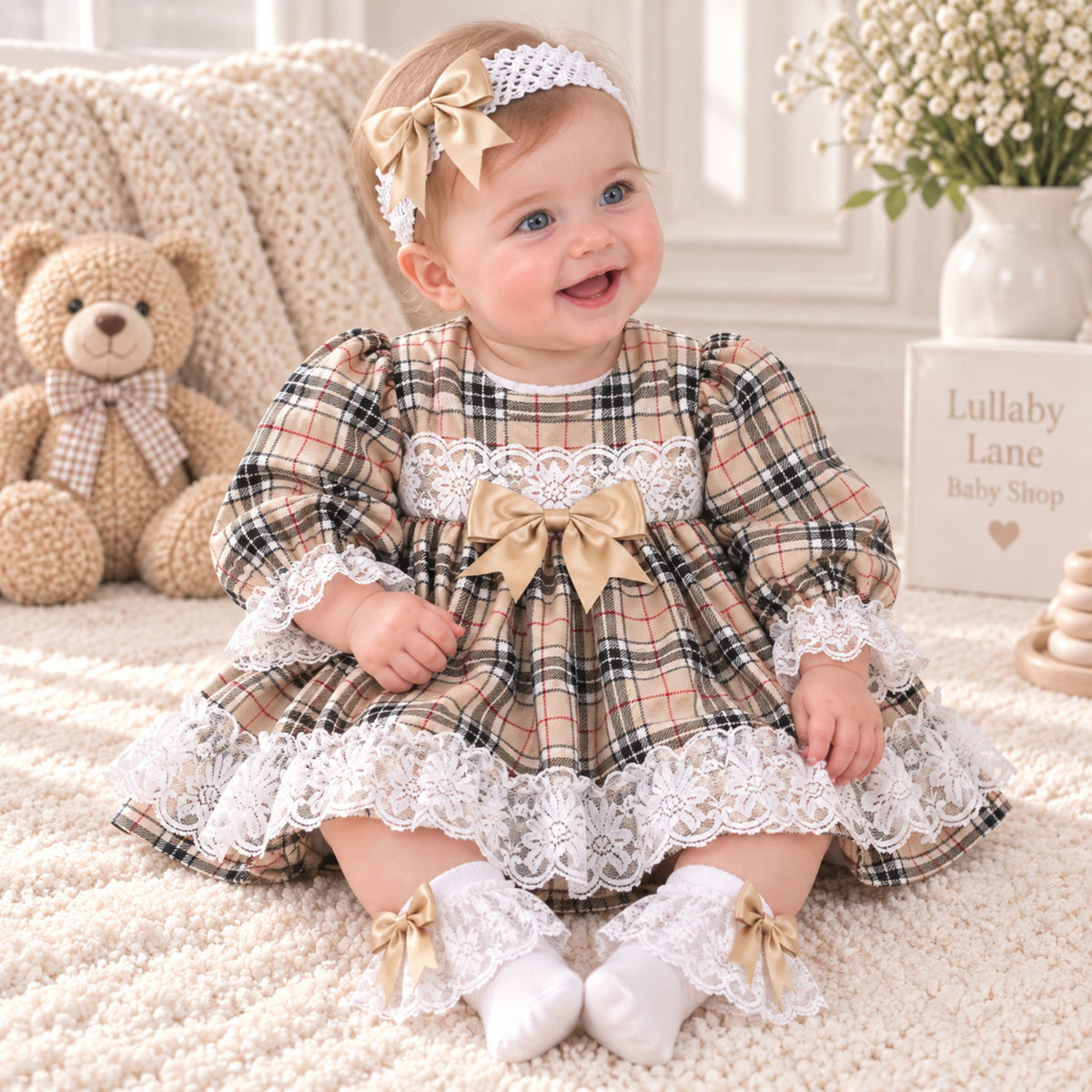 A baby girl sitting on a cream rug wearing a beige plaid dress with white lace trim, a matching headband, and white lace socks. A teddy bear and a sign reading 'Lullaby Lane Baby Shop' are visible in the background.