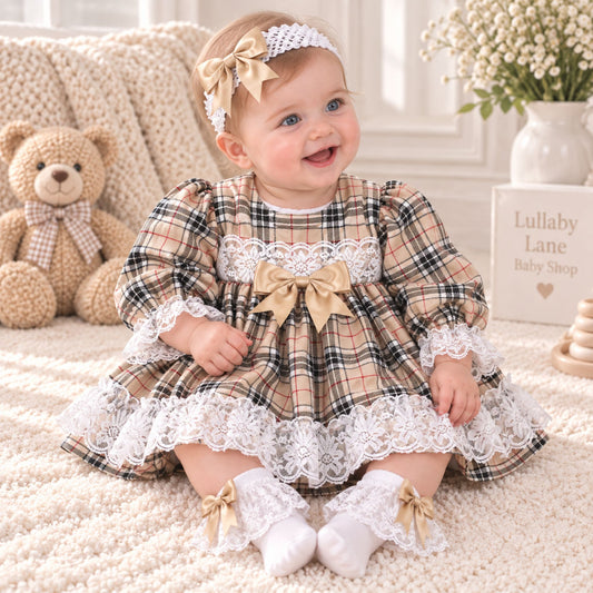 A baby girl sitting on a cream rug wearing a beige plaid dress with white lace trim, a matching headband, and white lace socks. A teddy bear and a sign reading 'Lullaby Lane Baby Shop' are visible in the background.