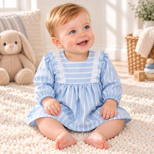A baby girl sitting on a white textured rug in a cozy nursery, wearing a light blue and white vertically striped long-sleeve dress with ruffled shoulder details.