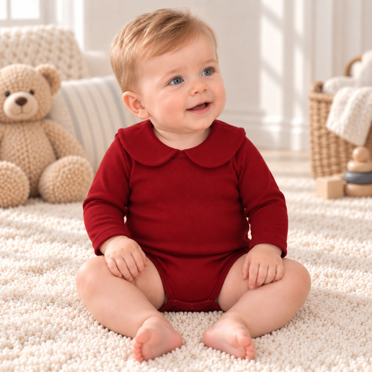 A baby sitting on a cream carpet wearing a long-sleeve red romper with a Peter Pan collar, with a teddy bear and toys in the background.