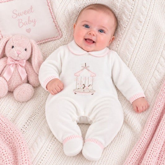 A baby lying on a cream knit blanket next to a pink plush bunny and a decorative pillow, wearing a white footed onesie with a pink giraffe-and-carousel print on the chest.