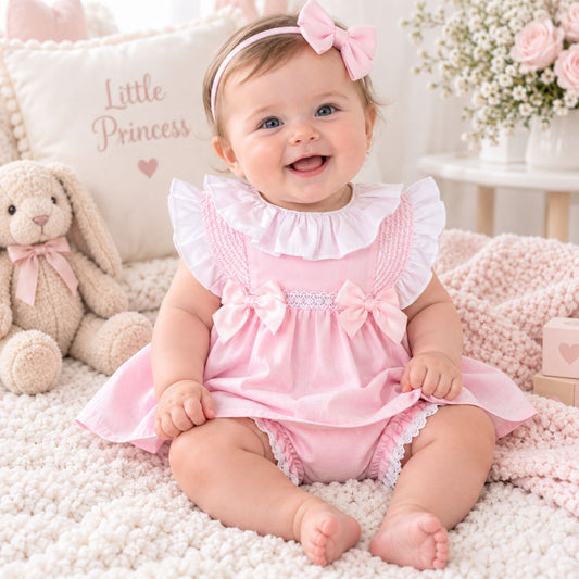 A baby girl sitting on a fluffy cream rug wearing a pink and white ruffled romper dress with matching pink bow headband. The background features a plush bunny toy, a decorative pillow that reads 'Little Princess', and soft pink flowers.