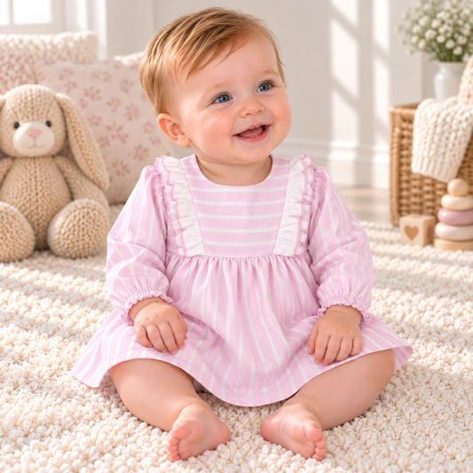 A baby girl sitting on a fluffy cream rug, wearing a pink and white striped long-sleeve dress with white ruffle trim on the chest. The background shows a cozy nursery setting with a plush bunny toy, pillows, and a basket.