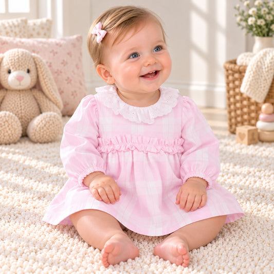 A baby girl sitting on a cream textured rug wearing a pink and white gingham long-sleeve dress with a white ruffled collar. The background features a cozy nursery setting with a plush bunny toy, patterned pillows, and a wicker basket.