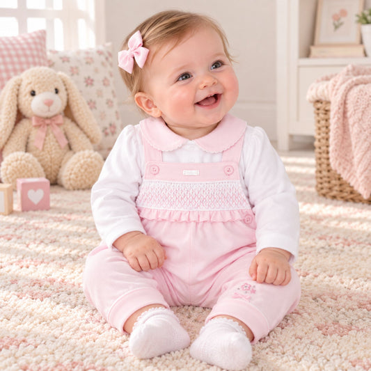 A baby girl sitting on a fluffy cream rug in a soft nursery setting, wearing a pink smocked overall romper over a white long-sleeve shirt. She has a pink bow in her hair, and a plush bunny toy is visible in the background.