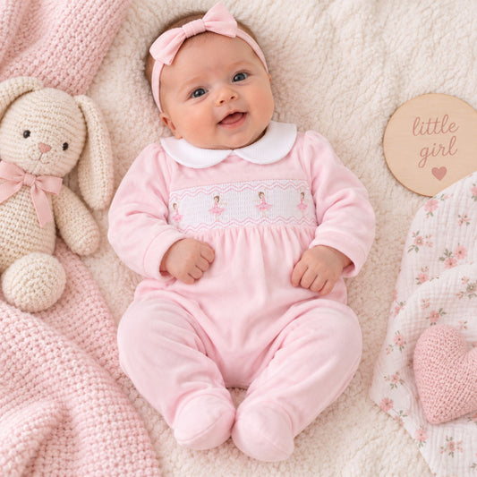 A baby lying on a soft cream blanket wearing a light pink footed onesie with a white Peter Pan collar and a pink bow headband. The scene includes a cream knitted bunny toy, a pink knit blanket, and a round wooden sign that reads 'little girl'.