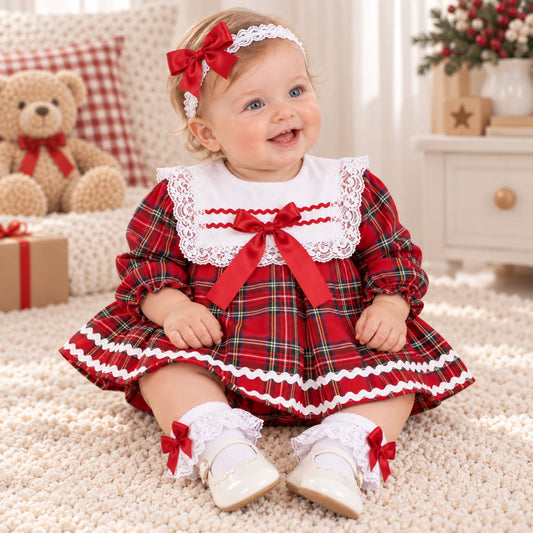 A baby girl wearing a festive red and green plaid dress with white lace trim, a red bow headband, and white shoes, sitting on a cream carpet in a decorated room.