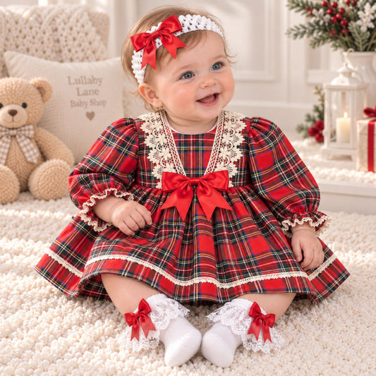 A baby girl wearing a festive red and green plaid dress with white lace trim, a matching red bow headband, and white socks with red bows, sitting on a fluffy cream rug in a decorated nursery.