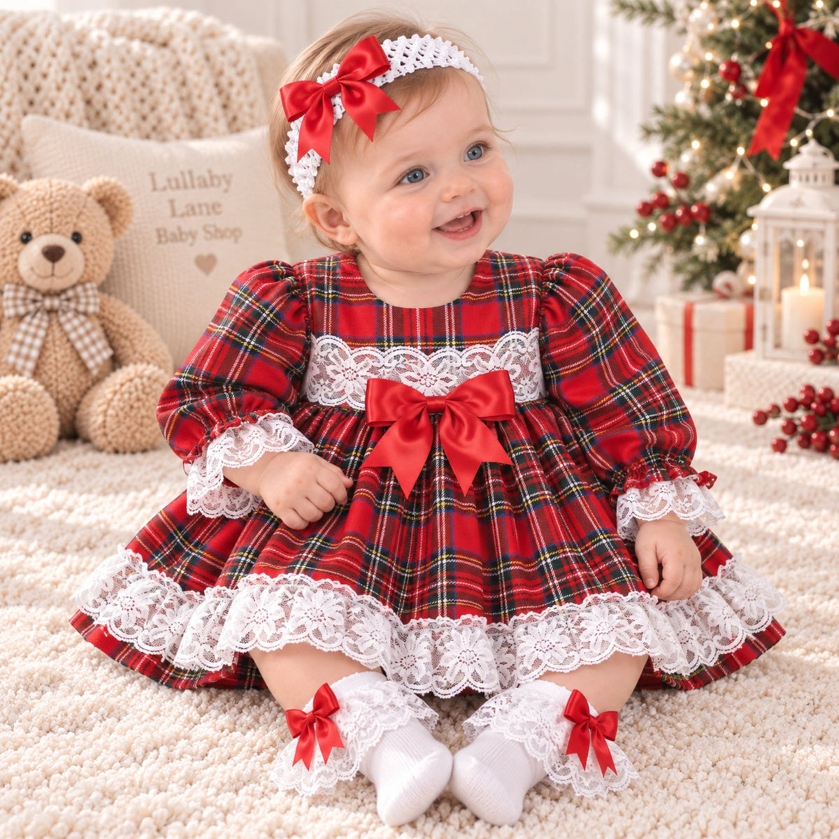 A baby girl wearing a festive red and green plaid dress with white lace trim, a matching red bow headband, and white socks with red bows, sitting on a cream rug in a decorated holiday room.