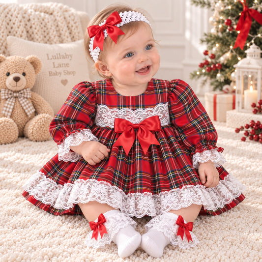 A baby girl wearing a festive red and green plaid dress with white lace trim, a matching red bow headband, and white socks with red bows, sitting on a cream rug in a decorated holiday room.