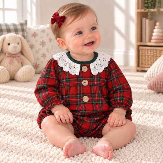 A baby girl sitting on a cream carpet in a cozy room, wearing a red and green plaid romper with a white lace Peter Pan collar and wooden buttons.