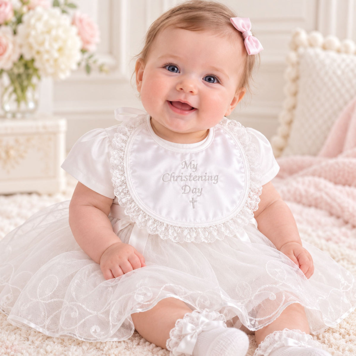 A baby girl wearing a white short-sleeve tulle christening dress with a lace-trimmed bib that reads 'My Christening Day' and a pink bow in her hair, sitting on a cream rug in a soft nursery setting.