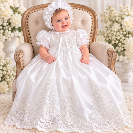 A baby wearing a white satin  christening gown dress with lace trim and a matching bonnet, sitting in a tufted cream armchair surrounded by white flowers.