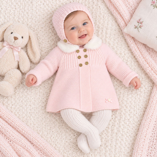 A baby wearing a light pink knit bonnet and matching cardigan with white faux-fur trim and gold buttons, lying on a cream knit blanket with a stuffed bunny beside them.