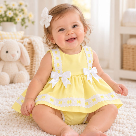 A smiling baby girl sitting on a white textured rug, wearing a yellow sleeveless dress with white lace trim and white bow details, along with a white bow in her hair.