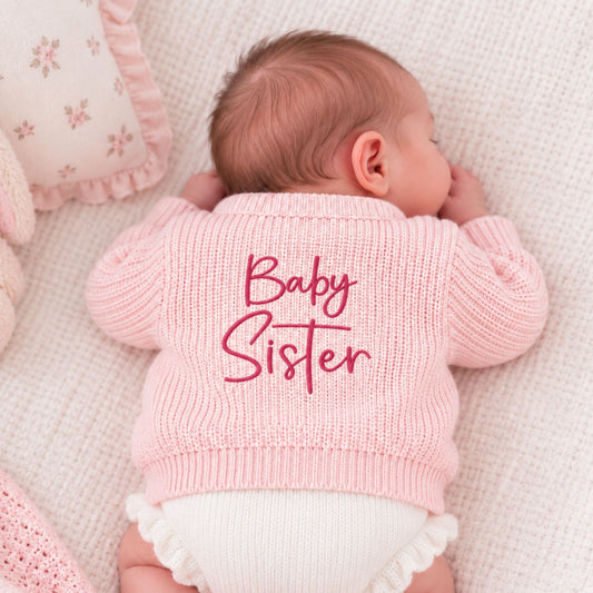 A sleeping newborn baby lying on their side, wearing a light pink ribbed knit sweater with 'Baby Sister' embroidered in dark pink script across the back. The baby is also wearing a white ruffled diaper.