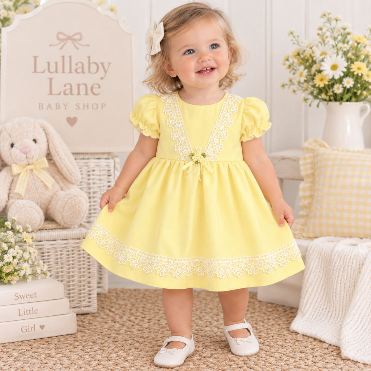 A toddler girl wearing a pale yellow dress with white floral lace trim and puff sleeves, standing in a styled nursery setting. The dress features a V-neckline with a small bow at the center and a matching lace hem.
