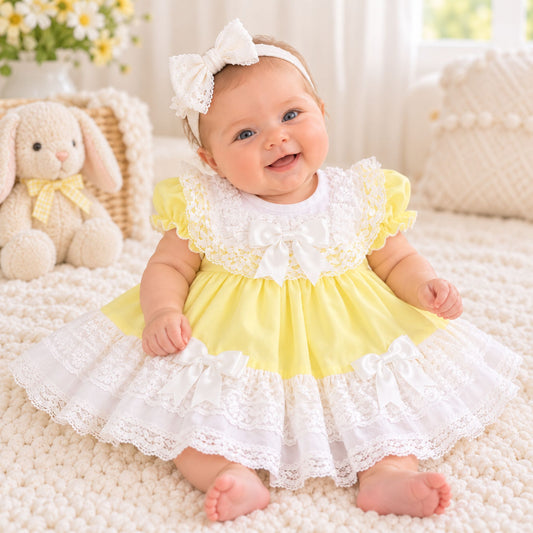 A baby girl wearing a yellow and white tiered dress with lace trim, a white bow headband, and a matching bow tie, sitting on a cream knit blanket next to a plush bunny toy.
