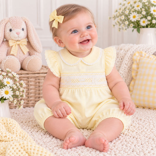 A baby girl sitting on a cream knit blanket, wearing a pale yellow smocked romper with ruffled sleeves and a matching yellow bow in her hair. The background features a plush bunny, daisy flowers, and a yellow gingham pillow.