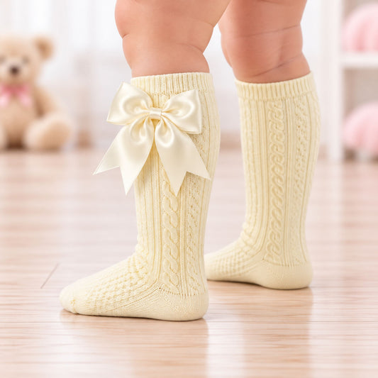 A close-up of a baby's legs wearing lemon-colored cable-knit socks with a large satin bow on the front, standing on a light wooden floor.
