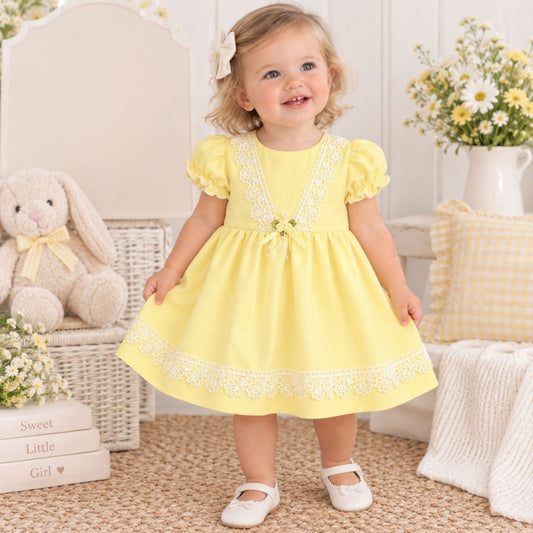 A toddler girl stands in a bright nursery wearing a pale yellow dress with white floral embroidery, puff sleeves, and a bow at the waist. The room includes a bunny plush, flowers, pillows, and a woven rug.
