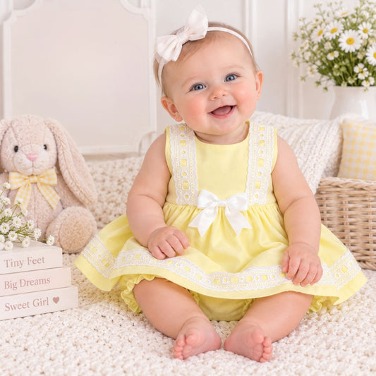 A baby girl sitting on a white textured rug, wearing a pale yellow sleeveless dress with white lace trim and a white bow headband. She is surrounded by a plush bunny toy, decorative books, and a basket with white daisies in a bright, soft setting.
