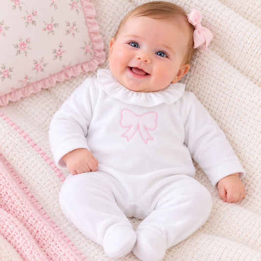 A smiling baby lying on a textured cream blanket with a pink floral pillow, wearing a white footed onesie with a pink bow on the chest and a pink hair bow.