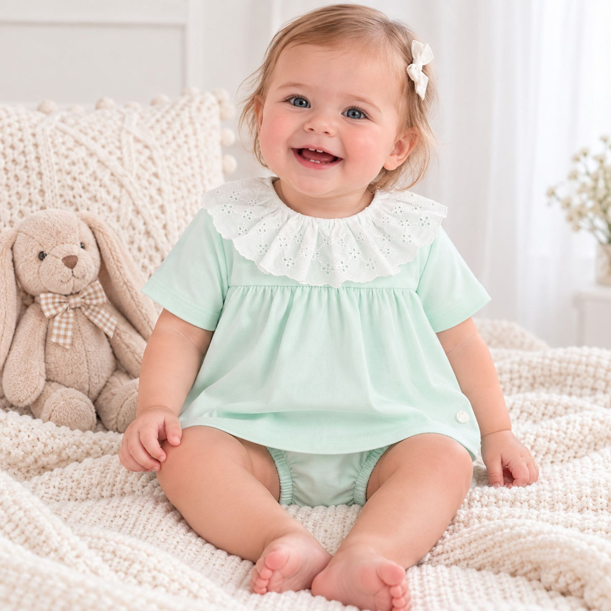 A baby girl sitting on a cream knit blanket, wearing a mint green short-sleeve romper with a white eyelet ruffled collar. She has a small white bow in her hair, and a plush bunny toy sits to her left.