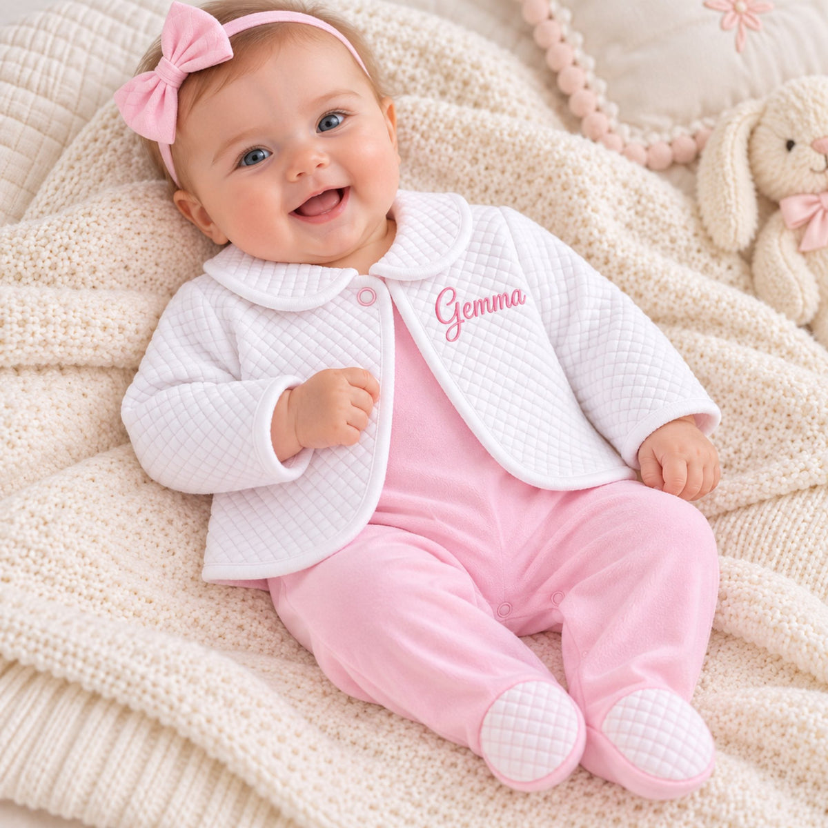 A smiling baby lying on a cream knit blanket, wearing a white quilted cardigan with pink piping and the name 'Gemma' embroidered on the chest, paired with pink footed onesie and a pink bow headband.