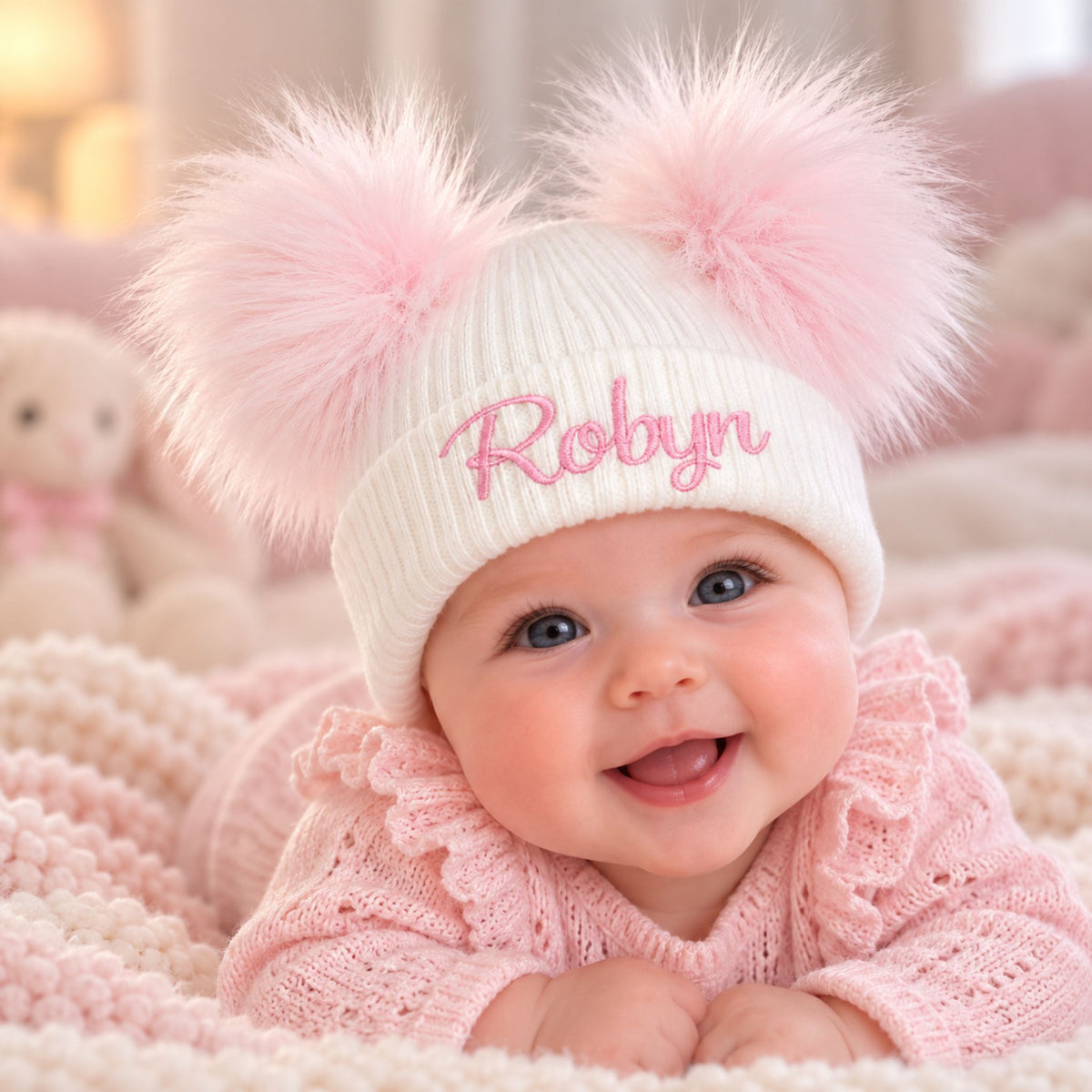 A smiling baby wearing a cream ribbed knit pom-pom hat with pink embroidered text reading 'Robyn' and fluffy pink pom-poms, lying on a soft pink blanket.