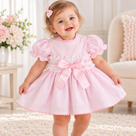 A toddler girl wearing a light pink baby dress with puffed sleeves, a ruffled waistband, and a large bow, standing in a bright, soft indoor room with flowers and a chair in the background.