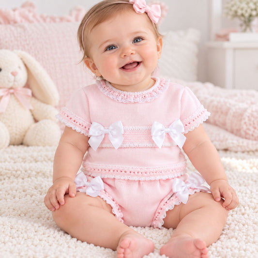 A smiling baby girl wearing a light pink short-sleeve romper with white lace trim and bow details, sitting on a white textured blanket in a soft nursery setting.