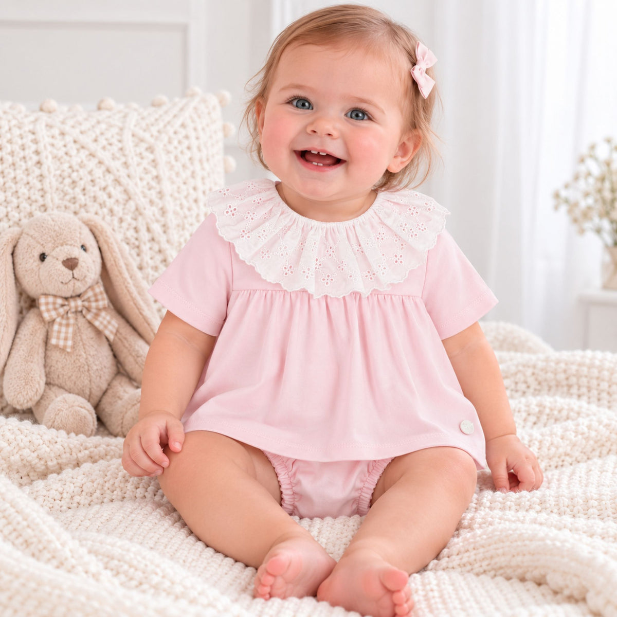 A baby girl sitting on a cream knit blanket next to a plush bunny, wearing a light pink short-sleeve dress with a white eyelet ruffled collar.